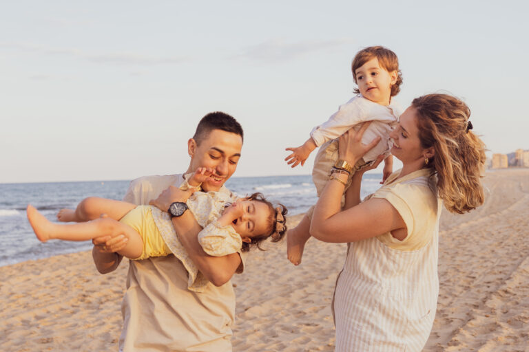 Sesión de fotos de familia en la playa: Padres jugando y levantando a los pequeños en un momento lleno de alegría y conexión.